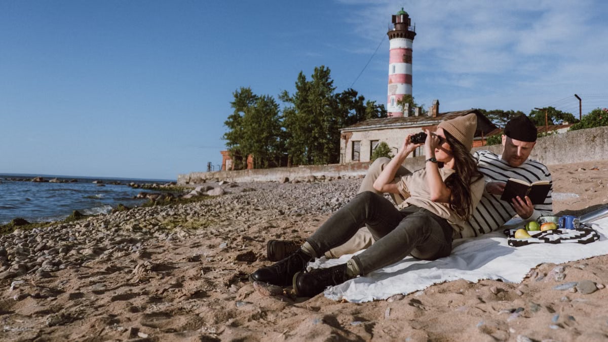 A couple enjoys a beach day by a lighthouse, with binoculars and a book for recreation.